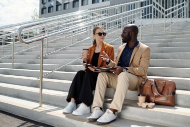 Genç, kendine güvenen bir borsacı kadın meslektaşına tabletle bir şey açıklarken ikisi de şehir ortamında merdivenlerde oturuyor.