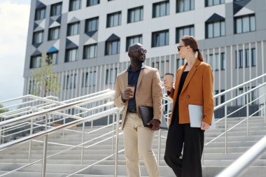 Lüks giysiler içinde modern genç çalışanlar birbirleriyle konuşuyor ve şehir ortamında aşağı kata taşınırken kahve içiyorlar.