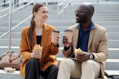Kültürler arası mutlu genç meslektaşlar kahve ve atıştırmalıklarla yemek arasında merdivenlerde otururken birbirlerine bakıyorlar.