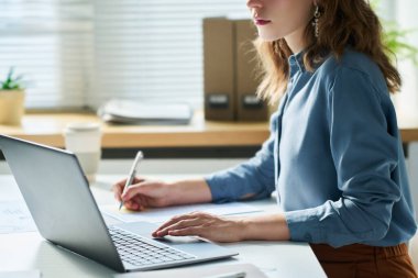 Mavi gömlekli, genç, beyaz yakalı kadın bir yandan dizüstü bilgisayarın önünde oturmuş, not alıyor ve online verileri analiz ediyor.