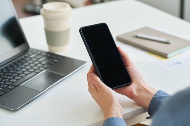 Ofiste çalışırken dizüstü bilgisayarı, kahve fincanı, not defteri ve kalemle genç bir iş kadınının elinde tuttuğu boş akıllı telefon ekranı.