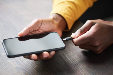 Genç Afrikalı Amerikalı adamın elleri kameranın önünde tahta masada otururken USB 'yi cep telefonuna bağlıyor.