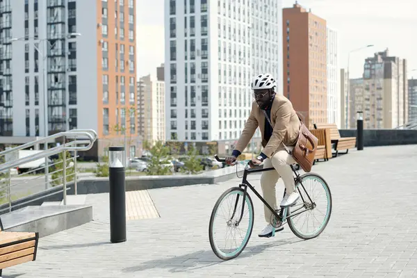 Young African American businessman with handbag on his neck cycling to work while moving along wooden bench against modern architecture