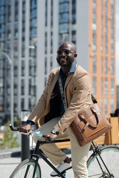 Young successful man in quiet luxury attire sitting on bicycle in front of camera and looking at you while riding to work in the morning