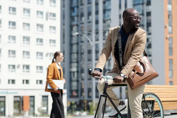 Young smiling businessman in quiet luxury apparel looking back over shoulder while sitting on bicycle and riding to work in the morning