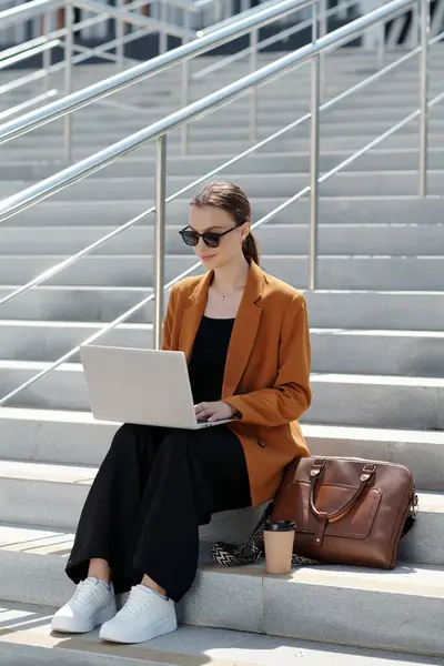 Young serious businesswoman in sunglasses and quiet luxury attire using laptop while sitting on staircase in urban environment and networking
