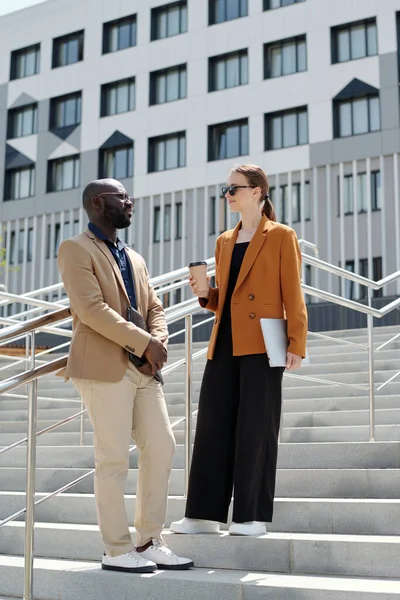 Two young intercultural employees in quiet luxury attire having coffee and chatting at break while standing on staircase in urban environment