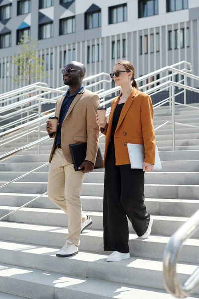 Two young smiling colleagues in formalwear carrying cups of coffee and gadgets while walking downstairs against modern architecture