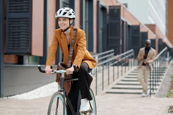 Happy young female employee in quiet luxury attire and safety helmet riding bicycle in the morning against male broker using smartphone