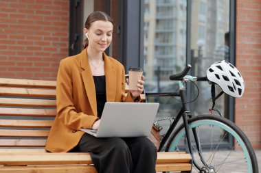 Elinde bir fincan kahveyle bankta oturup dışarıda dizüstü bilgisayara bakarken online video izleyen genç ve aktif bir iş kadını.