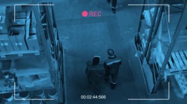 Yüksek açılı CCTV görüntülerinde etnik çeşitliliğe sahip kadın ve erkekler depoda yürürken planlar tartışıyorlar ve palet kriko çekiyorlar.