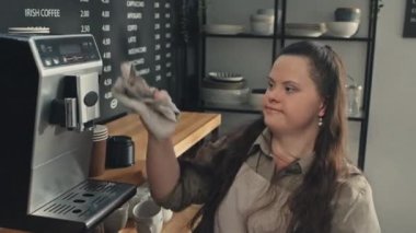 Down sendromlu genç bayan barista kafe temizleme makinesinde çalışıyor ve onu kuru havluyla siliyor.