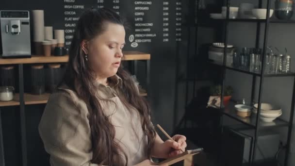 患有唐氏综合症的年轻女服务员站在柜台后面 手里拿着为顾客准备的咖啡杯和甜甜圈 在笔记本上记笔记 — 图库视频影像