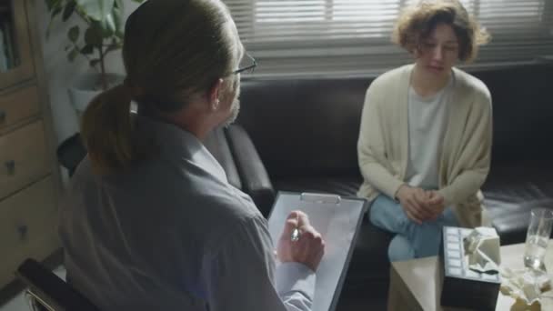 治療セッション中にメモを作る患者の前に座っているプロの精神保健専門家の高角度オーバーショット — ストック動画