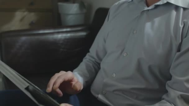 デジタルタブレットで何かをスクロールオフィスのソファに座って顔にひげを持つ成熟した白人男性のミディアムクローズアップ傾斜ショット — ストック動画