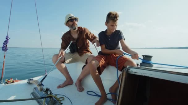 成熟したコーカサス人の男は彼の十代の息子にウィンチとロープを使用してヨットの帆を操作する方法を教えています — ストック動画