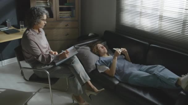 年轻女子躺在皮革沙发上的高透视告诉专业精神治疗师她的抑郁症 — 图库视频影像