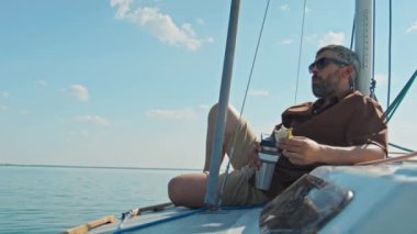 Beyaz saçlı ve sakallı beyaz bir adam yaz günü yatta oturmuş sandviç yiyor, çay içiyor ya da kahve içiyor.