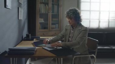 Zarif, olgun, beyaz kadın psikoterapist masada oturuyor. Elinde kağıtlarla dizüstü bilgisayarını açıyor.