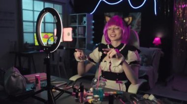 Genç Cosplayer odasında oturmuş, akıllı telefon kamerasıyla video kaydederken takipçilerine makyaj ürünleri gösteren blog içeriği yaratıyor.