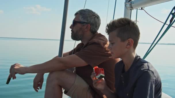 顔にひげを被った白人男性と10代の息子は スイカを食べながら何かをしゃべっているヨットに乗ってリラックスしました — ストック動画