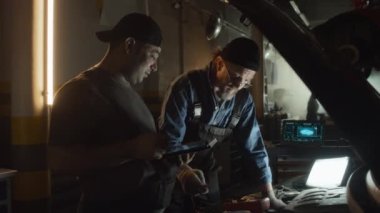 Otomobil tamirhanesinde çalışan olgun, beyaz ve genç Orta Doğulu erkekler otomobil motorunun işlevselliğini kontrol ediyor.