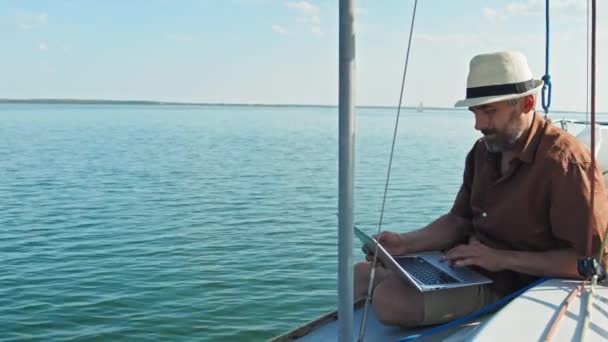 成熟したコーカサス人の男は ノートパソコンで何かをテキストするヨットの夏の日に顔にひげを付ける — ストック動画