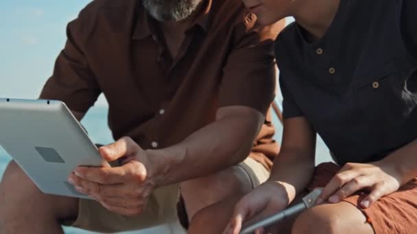 デジタルタブレットの写真を見ているボートに座っているひげ付き男と10代の少年の中央セクションの作物ショット — ストック動画