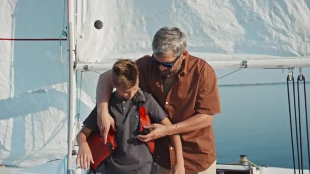 中镜头的成熟白人男子站在码头上帮助他的少年儿子穿上救生衣 — 图库视频影像
