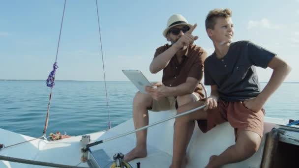 成熟的男人和他十几岁的儿子在一起度过了夏天 他们一起乘游艇在数字平板电脑上观看一些东西 然后遥望远方 — 图库视频影像
