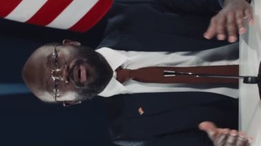 ABD, Almanya ve Avrupa Birliği bayraklarıyla uluslararası zirvede masada oturan ve izleyicilere açılış konuşması yapan orta yaşlı Afrikalı Amerikalı politikacının orta yaş çekimi.