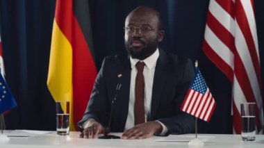 Afrika kökenli Amerikalı erkek hükümet yetkilisinin mikrofon önünde İngiltere, ABD, AB ve Alman bayraklarıyla oturduğu orta boy bir fotoğraf ve uluslararası konferans sırasında konuşma yapması.