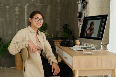 Esmer kadın fotoğrafçı bilgisayar monitörüyle masada oturuyor. Stüdyoda çalışırken genç modelin yeni görüntüleri ekrana yansıyor.