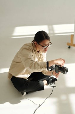 Genç profesyonel fotoğrafçı stüdyonun zemininde otururken çekim yapmadan önce fotoğraf makinesinin ayarlarını ayarlıyor.