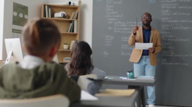 Erkek, neşeli, Afro-Amerikan öğretmenin sınıfın tahtası önünde durup gündüz cevap veren öğrenciyi dinlediği bir sahne.