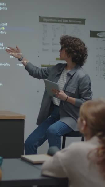 黒板の前で椅子に座ってスクリーンプロジェクションを指して説明する女性のカウシアン教師のフルショット — ストック動画