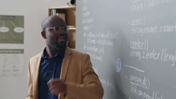教室の黒板の前に立っている男性アフリカ系アメリカ人教師のサイドビューメディアのクローズアップ 昼間の学生にプログラミング教材の説明 — ストック動画