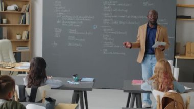 Erkek Afro-Amerikan öğretmenin okul sınıfında karatahtanın önünde durup gündüz programları üzerine konferans vermesi.