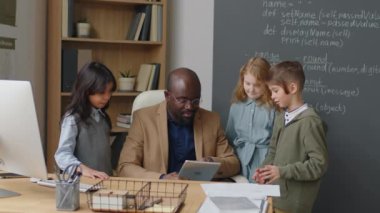 Etnik çeşitlilikte üç ilkokul öğrencisinin erkek Afrikalı Amerikan öğretmeninin etrafında durup gündüz vakti sınıfta tartışmalarının orta boy fotoğrafı.