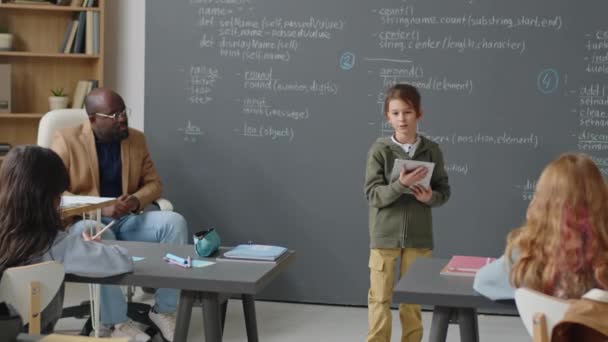 10歳のコーカサス人の少年がタブレットを握るクラスの前に立ってレポートを作り 机に座っているアフリカ系アメリカ人教師が彼に耳を傾けている — ストック動画