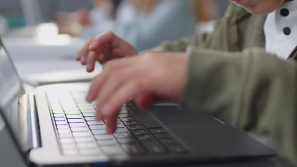 ノートパソコンのキーボードに何かを入力する学校の教室で机に座っている小学生の手に焦点を当てる — ストック動画