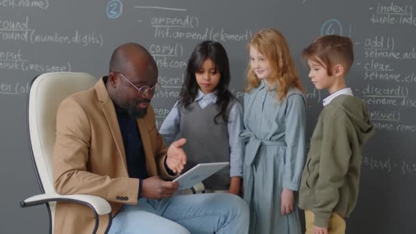 坐在黑板前的非洲裔美国男教师在课堂上向3名不同种族的小学生讲解主题的中等全景照片 — 图库视频影像
