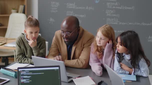教室の机に座ってノートパソコンを見て 近くに立っている民族的に多様な小学生に何かを説明する男性のアフリカ系アメリカ人教師のミディアムショット — ストック動画