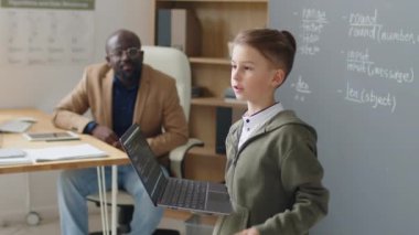 Orta boy beyaz çocuk okulda karatahtanın önünde dizüstü bilgisayarla duruyor ve gündüz programlama dersleri üzerine proje sunuyor.