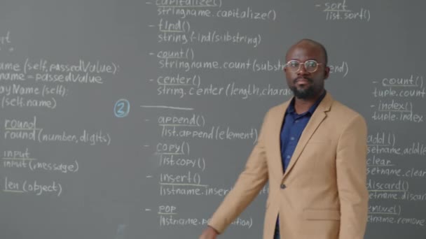 男性のアフリカ系アメリカ人の教師の中型肖像画が 昼間のプログラミング教室でカメラを撮るためにブラックボードにやってくる — ストック動画