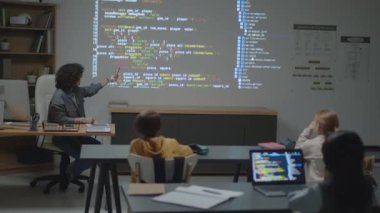 Kafkas kadın öğretmenin sınıfta oturduğu tam bir fotoğraf. Ekran projeksiyonunu işaret ediyor. İlkokul öğrencilerine şifreli bir şekilde anlatıyor.