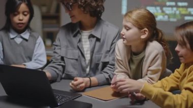 Beyaz kadın öğretmen ve üç farklı ilkokul öğrencisinin yan yana oturup dizüstü bilgisayara bakmaları ve programlama dersi sırasında tartışmaları.