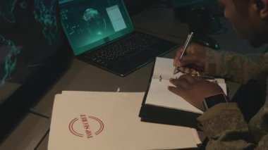 Yüksek açılı, odaklanmış Afrika kökenli Amerikan askeri gözetim memurunun gece vakti elinde haritayla bilgisayar ekranına bakıp notlar alırken görüntüsü.