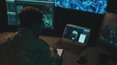 Erkek Afro-Amerikan askerin bilgisayar ekranının önünde oturup deftere bir şeyler yazarken omuz üstünden çekilen fotoğrafı.