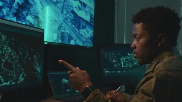 男性アフリカ系アメリカ人の軍事監視官がデスクに座ってコンピュータ画面を観察し プログラムを分析してメモを作成する中型ショット — ストック動画